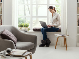 Young woman sitting next to a window and connecting with her laptop