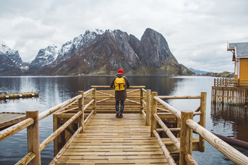 Traveler man with a yellow backpack wearing a red hat standing on the background of mountain and lake wooden pier. Travel lifestyle concept. Shoot from the back