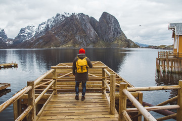 Fototapeta na wymiar Traveler man with a yellow backpack wearing a red hat standing on the background of mountain and lake wooden pier. Travel lifestyle concept. Shoot from the back