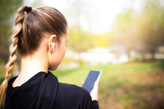 Young Smiling Girl Making Sport And Running In The Park Using Her Phone To Listen The Music With Wireless Headphones On Sunset In The City Watching The Screen