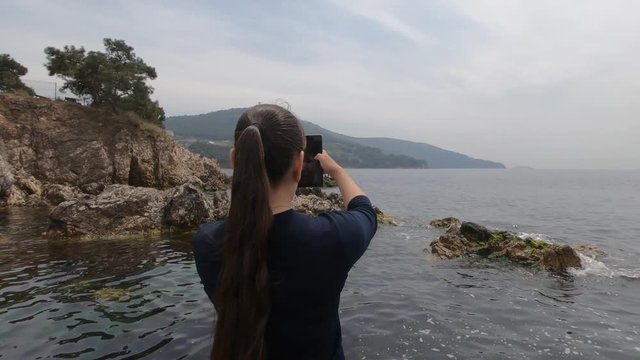 A girl of Asian appearance stands on a rocky beach in Turkey and takes pictures of beautiful landsc