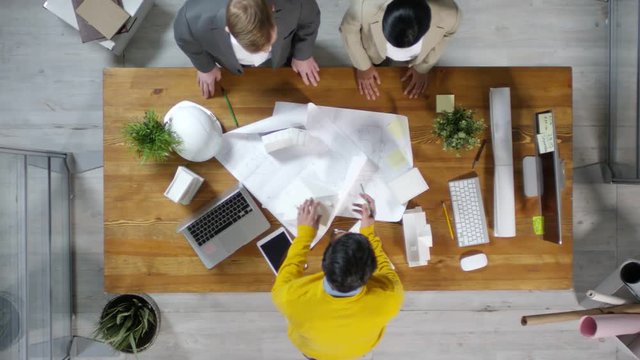 Directly above view of businessman and businesswoman standing at wooden table and talking to mixed-race architect standing in front of them and drafting on blueprint
