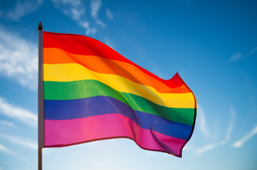 Gay pride rainbow flag fluttering backlit in the sun against soft blue sky copy space