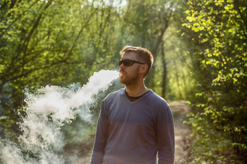 Vape man. An adult white bearded man in glasses smokes an electronic cigarette outside in the forest in sunny day. Bad habit that is harmful to health.