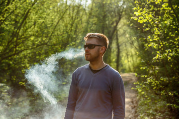 Vape man. An adult white bearded man in glasses smokes an electronic cigarette outside in the forest in sunny day. Bad habit that is harmful to health.