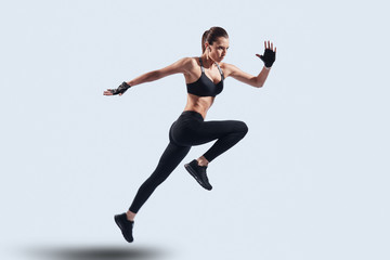 Fototapeta na wymiar Full of energy. Full length of attractive young woman in sports clothing jumping while hovering against grey background
