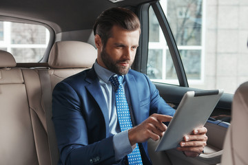 Using modern technologies. Young confident and stylish businessman in formal wear is working on digital tablet while sitting on the back seat of his car.