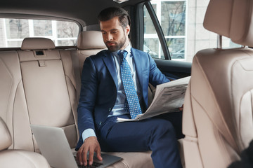 Working from car. Young confident businessman in formal wear is working on laptop and reading fresh newspaper while sitting on the back seat of his car.