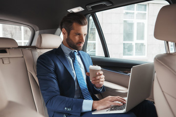Focused on work. Busy young businessman in formal wear working on laptop and drinking hot coffee while sitting in his car.