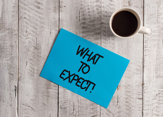 Text sign showing What To Expect. Business photo showcasing asking about regard something as likely to happen occur Pastel Colour paper placed next to a cup of coffee above the wooden table