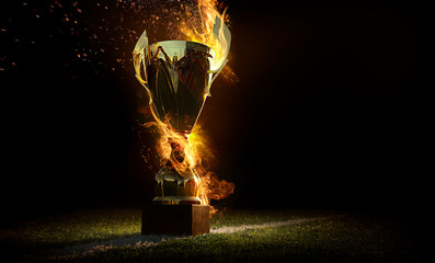 Sports background. Burning trophy goblet. Winner in a competition. Fire and energy. Football field with golden goblet. - obrazy, fototapety, plakaty
