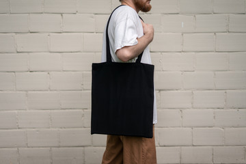 Urban mockup of tote bag. Men holding black cotton tote bag on a brick wall background. Template...