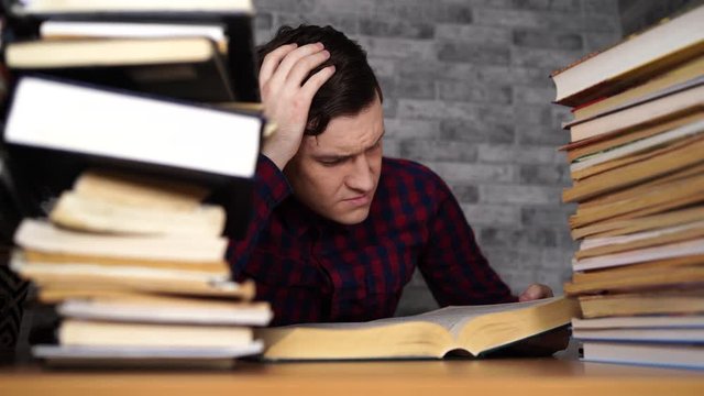 Man student boring reading book at library with a lot of books in university. Student disheartened reading book for examination.