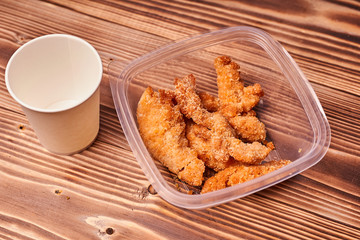 Chicken strips in a plastic container with a paper Cup on a wooden table.