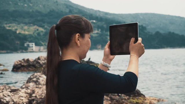 The girl stands on a rocky beach and takes pictures on the tablet beautiful landscape of the sea and green hills