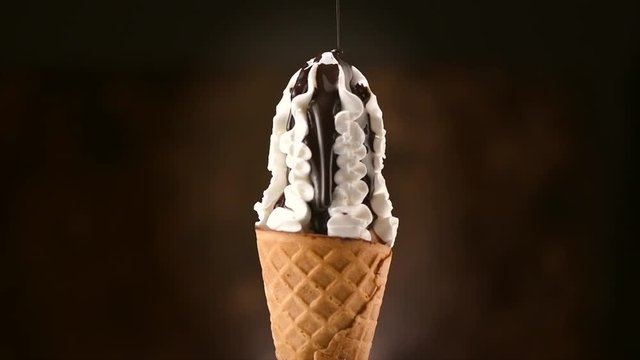 Ice cream cone close-up. Pouring chocolate syrup topping on Icecream in waffle cone, rotating on black background. 4K UHD video footage. 3840X2160