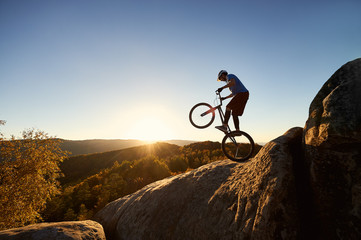 Fearless cyclist standing on back wheel on trial bicycle. Professional sportsman rider balancing on the edge of big boulder on the top of mountain at sunset. Concept of extreme sport
