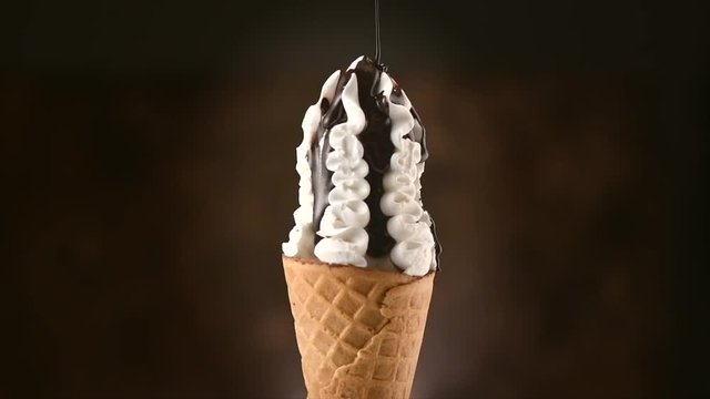 Ice cream cone close-up. Pouring chocolate syrup topping on Icecream in waffle cone, rotating on black background. 4K UHD video footage. 3840X2160