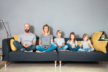 Mother and father practicing yoga poses with their three children.Whole family sitting in lotus position on sofa at home