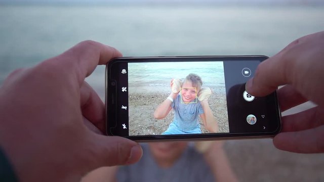 Closeup point of view shoot of male hands holding mobile smartphone and taking photoes of kut smiling happy kid holding two sea shells. Father photographing his son at summer beach during vacations.