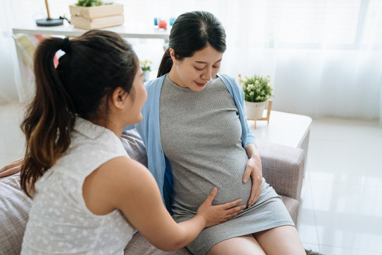 elegant beautiful asian korean woman expecting first baby sitting on couch in living room at home. two ladies best friends together looking at big belly future motherhood prepared concept. girl touch