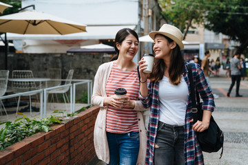 Young asian female friends walking around small town in russia. happy girls traveler talking and carry coffee to go in hands shopping relax in outdoor holidays market on sunny day in spring.