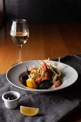 Seafood made dish with king prawns, mussels, squid, greens and citrus served with white wine and black olives on a white plate and wooden table