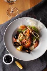 Seafood made dish with king prawns, mussels, squid, greens and citrus served with white wine and black olives on a white plate and wooden table