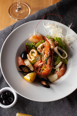 Seafood made dish with king prawns, mussels, squid, greens and citrus served with white wine and black olives on a white plate and wooden table