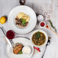 Top view of a three course set menu: beef stroganoff with mashed potatoes, roasted crunchy cutlet with rice and hot sorrel soup, served on a white wooden table