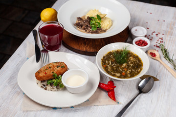Top view of a three course set menu: beef stroganoff with mashed potatoes, roasted crunchy cutlet with rice and hot sorrel soup, served on a white wooden table