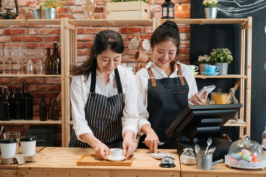 two girl barista and cashier in aprons standing and talking behind bar counter in cafe. waitress counting bills on calculator while holding notebook. smiling woman staff preparing coffee for customer