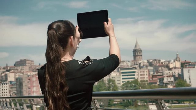 Girl tourist stands near a beautiful area of the city and takes pictures on the tablet blue sky