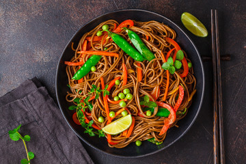 Vegan buckwheat soba noodles with vegetables in black plate on dark background, top view.