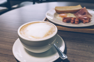 White cup of coffee on wooden table with breakfast set in vintage tone 