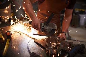 Close-up of unrecognizable workman in apron standing at metal table and using tool with grinding disc while cutting metal bar 