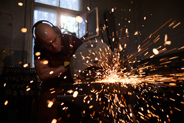 Serious concentrated young blacksmith in ear protectors and protective goggles using steel cutting...
