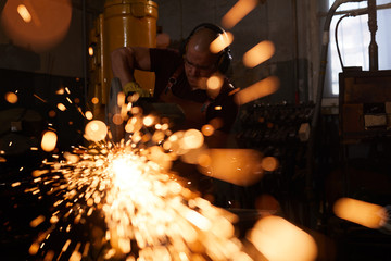 Busy factory worker in ear protectors standing in workshop and sawing metal with cutting machine