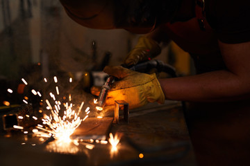 Close-up of unrecognizable laborer in blacksmith gloves bending while welding metal piece with...
