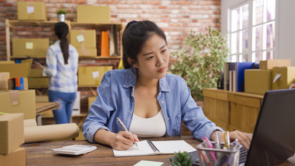 Young casual beautiful women entrepreneur sitting at work desk using laptop computer writing. two female teenager business owner partner colleague office many stuff to do. parcels boxes in workplace