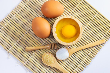 Top view raw egg in wooden bowl with salt and sugar in wooden spoon on bamboo mat