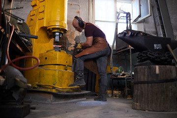 Concentrated young man in ear protectors and leather apron fixing metal piece in forging press machine