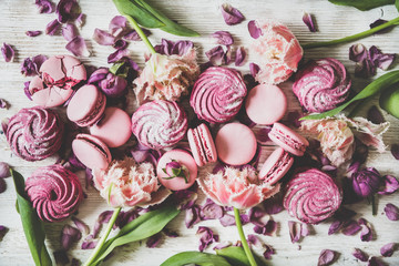 Flat-lay of sweet pink macaron cookies, lilac marshmallows and spring flowers over wooden background, top view. Food texture, background and wallpaper