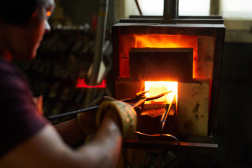 Over shoulder view of busy experienced blacksmith using tongs while putting metal piece into heated...