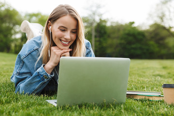 Photo of gorgeous caucasian woman smiling and using laptop with earphones while lying on green grass in park