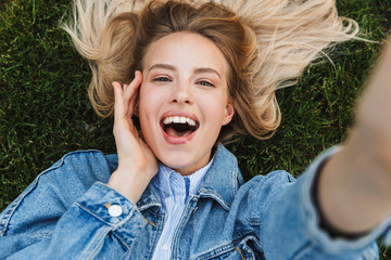 Photo of blonde kind woman taking selfie photo with hand on her face while lying green grass in park