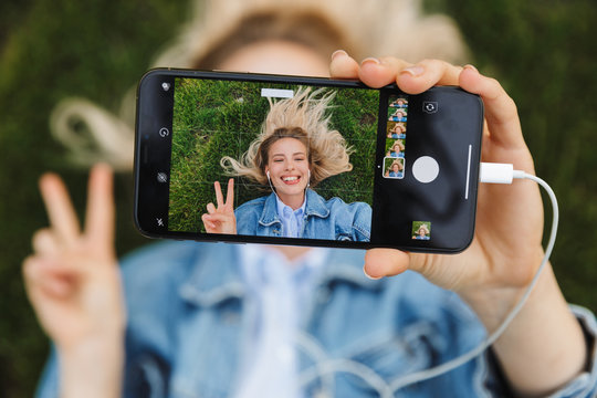 Photo of blonde happy woman taking selfie photo on cellphone and gesturing peace sing while lying green grass in park