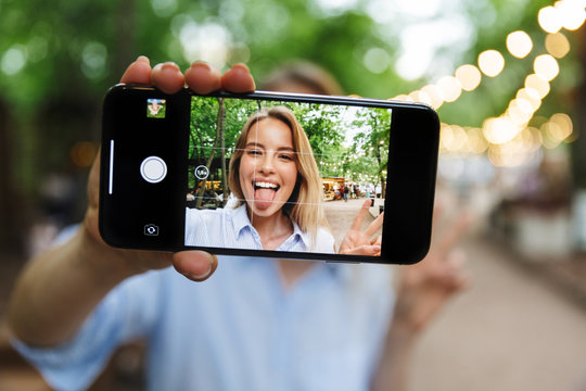 Photo of blonde smiling woman selfie photo on smartphone with sticking out her tongue and gesturing peace sing