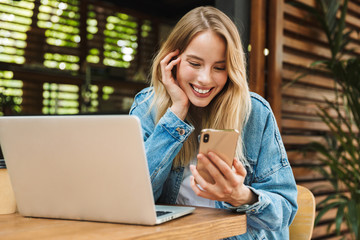 Portrait of blonde smiling woman using laptop and cellphone in cafe outdoors