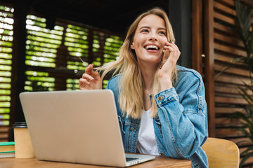 Portrait of caucasian laughing woman talking on cellphone while using laptop in cafe outdoors